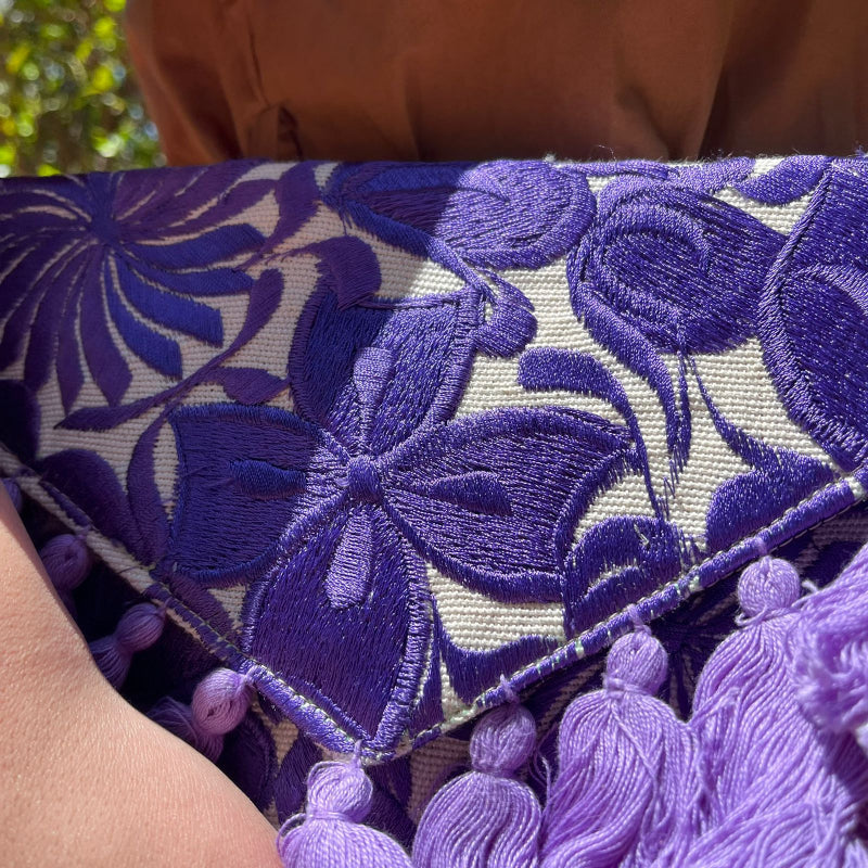 Purple floral embroidered clutch bag with tassels held by a person wearing a brown dress outdoors