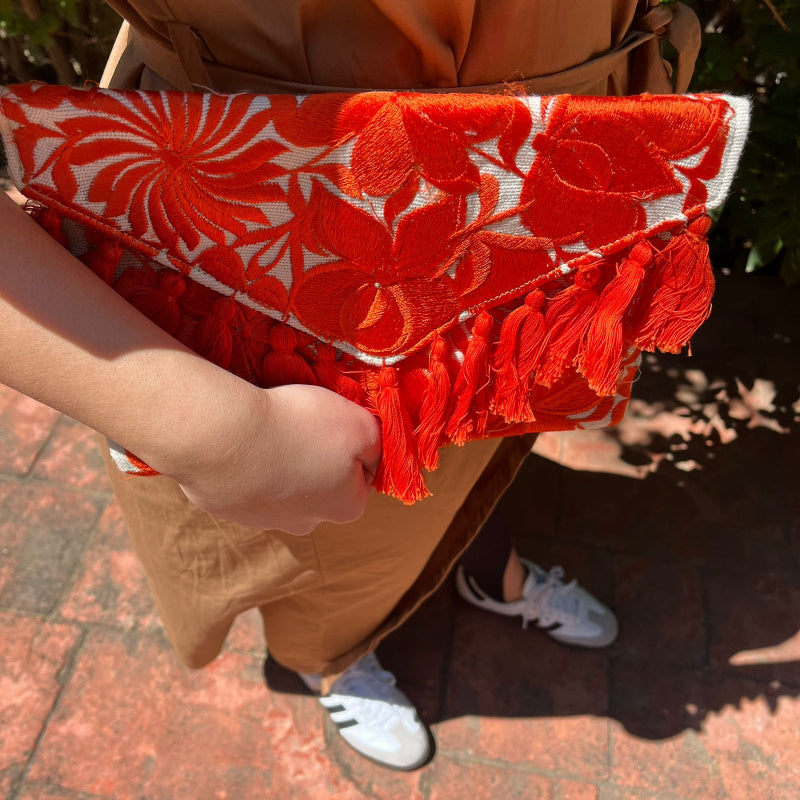 Red floral embroidered clutch bag with tassels held by a person wearing a brown dress outdoors