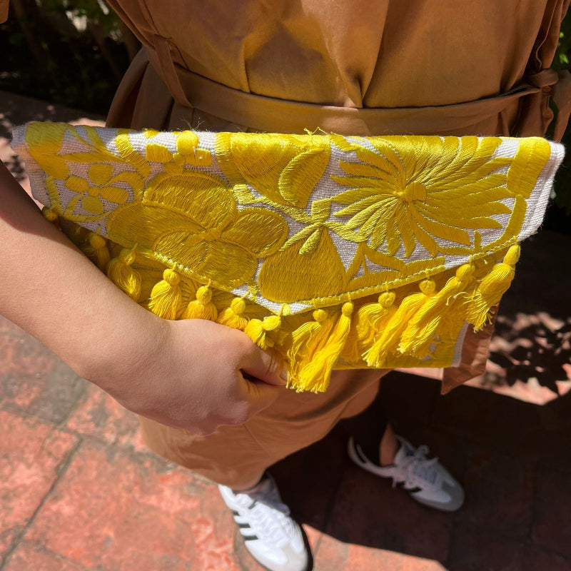 Bright yellow floral embroidered clutch bag with tassels held by a person wearing a brown dress outdoors