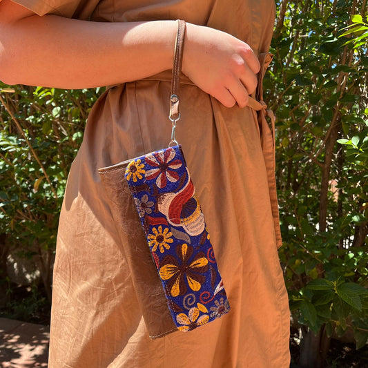 Hand holding a handmade brown leather clutch with colorful floral embroidery and wrist strap outdoors