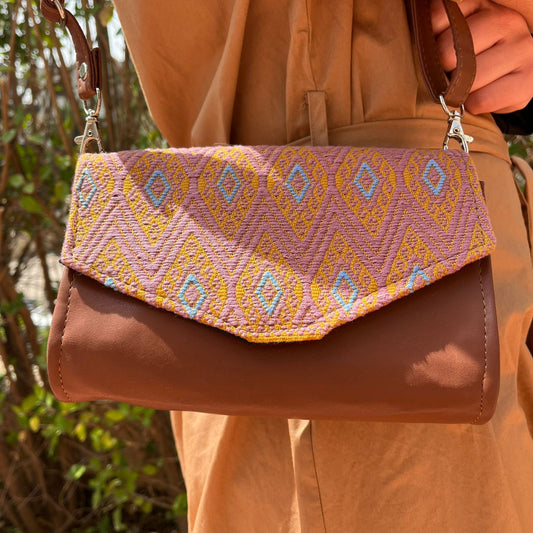 Close-up of a brown leather crossbody bag with mauve and gold geometric woven flap worn outdoors