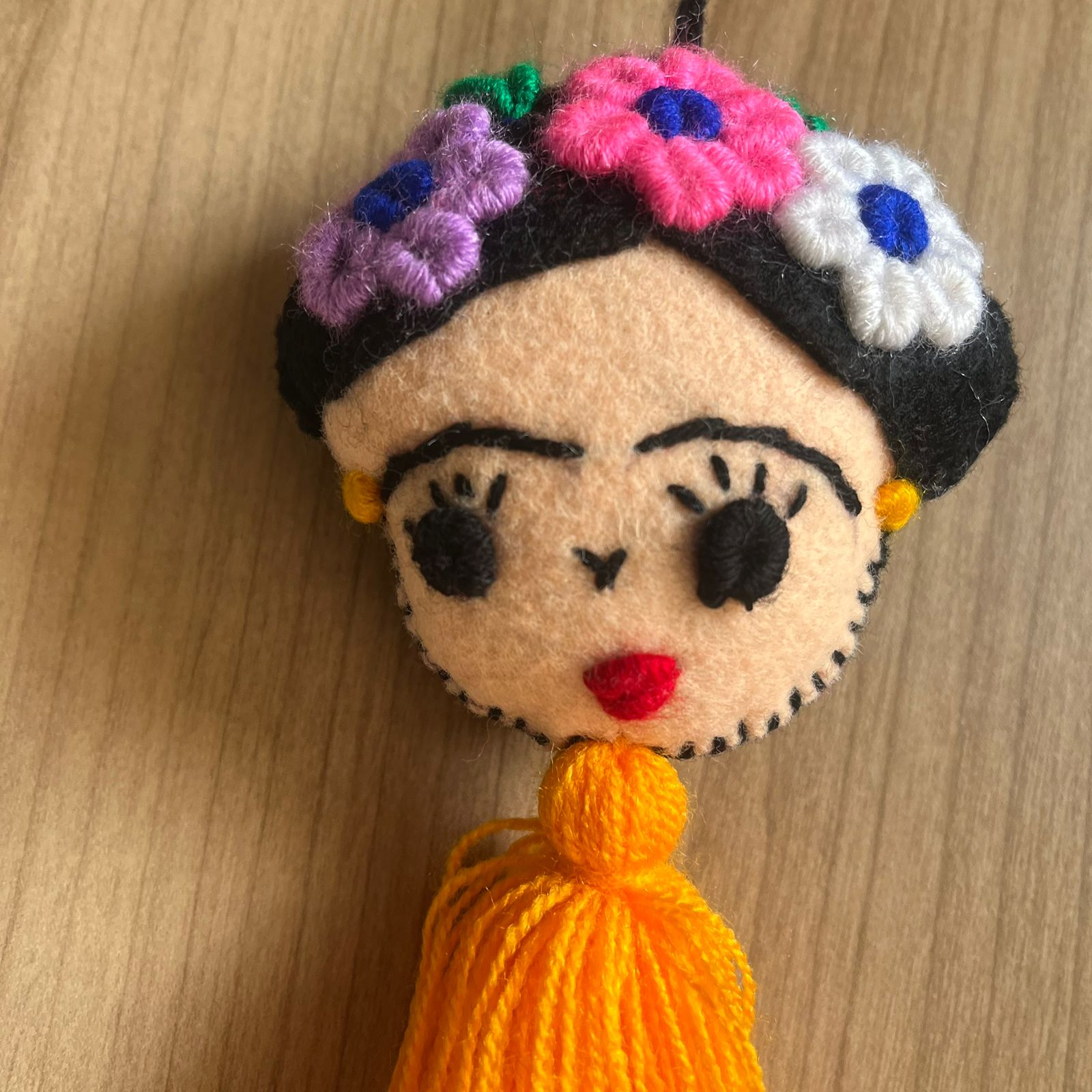 Handmade Frida Kahlo felt ornament with multicolor floral embroidery and bright orange tassel hanging on wooden background
