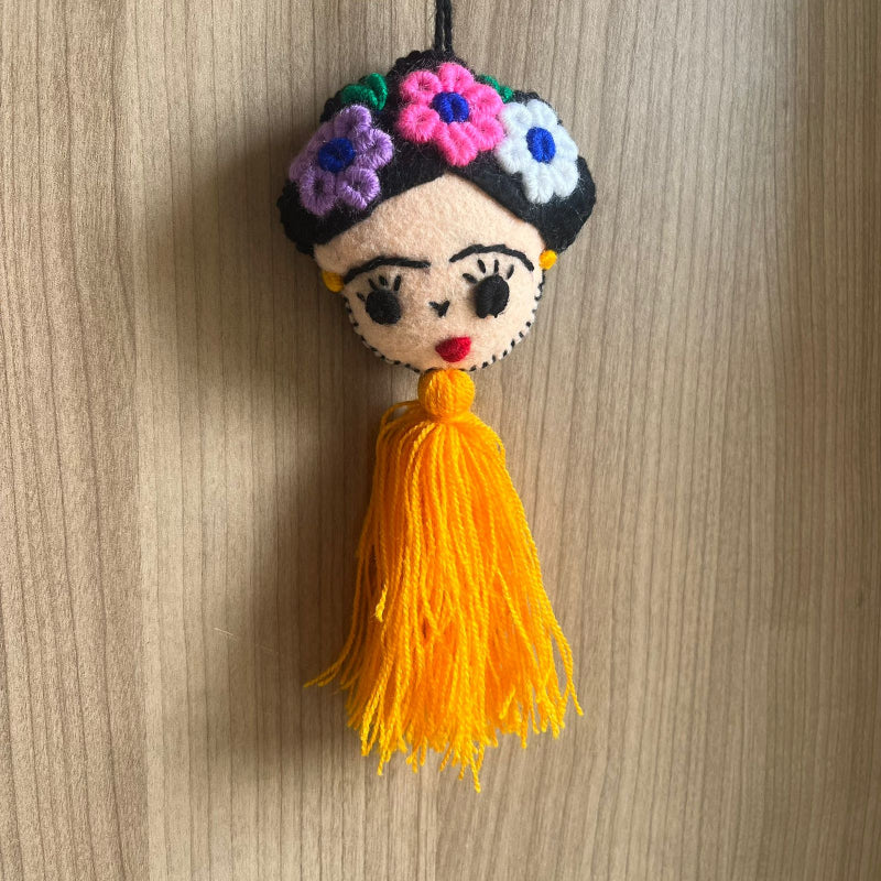 Handmade Frida Kahlo felt ornament with multicolor floral embroidery and bright orange tassel hanging on wooden background