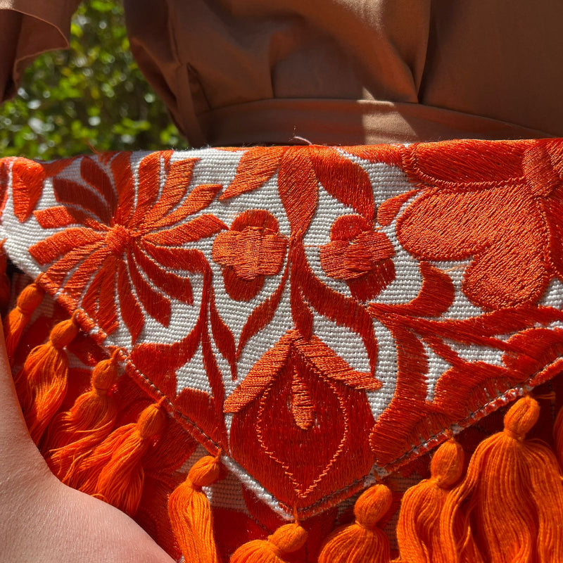 Bright orange floral embroidered clutch bag with tassels held by a person wearing a brown dress outdoors