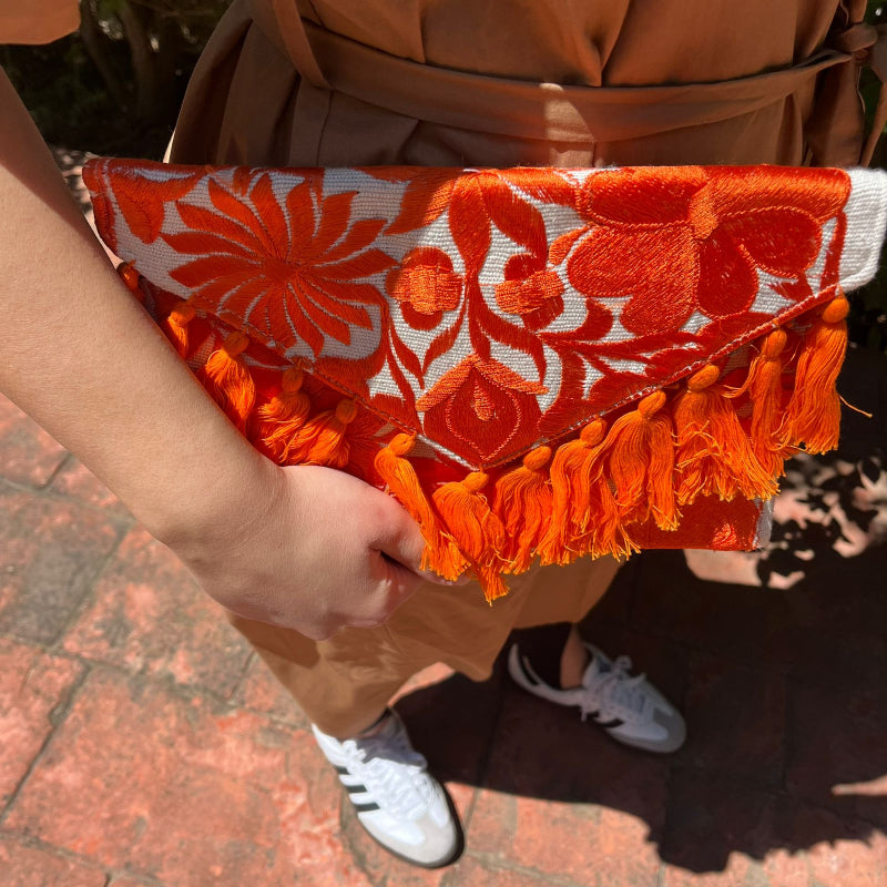 Bright orange floral embroidered clutch bag with tassels held by a person wearing a brown dress outdoors