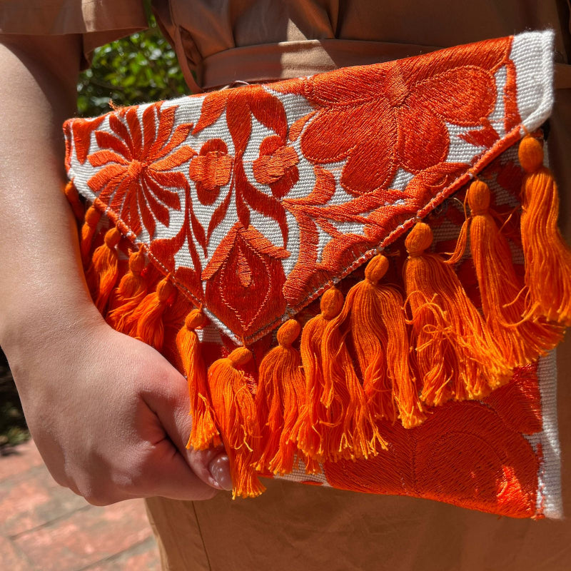 Bright orange floral embroidered clutch bag with tassels held by a person wearing a brown dress outdoors