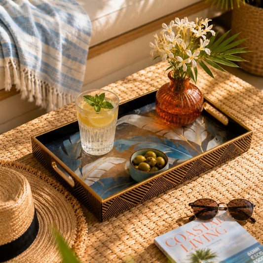 A styled outdoor lounge scene with a woven rattan tray holding a glass of lemonade, olives, and a small vase of white flowers, placed on a woven rug beside a straw hat, sunglasses, and a magazine. A cream sofa with a blue pillow and striped throw is in the background, along with palm leaves and a potted plant.