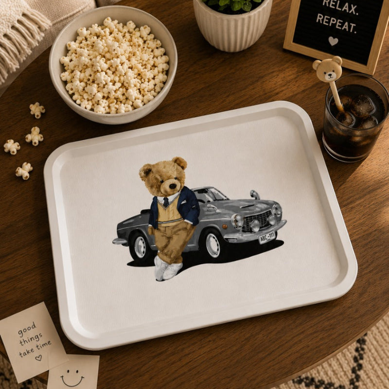 A white rectangular tray with rounded corners sits on a wooden table, printed with an illustration of a teddy bear standing next to a black vintage car. The tray is styled in a cozy room setting with a bowl of popcorn, a small potted plant, a flower-shaped throw pillow, and a sign that reads 'PAUSE, SMOKE, RELAX, REPEAT.