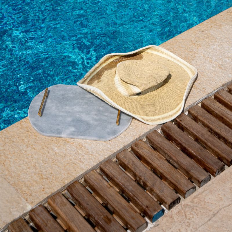 Light grey oval capsule-shaped marble tray with brass handles placed by the pool