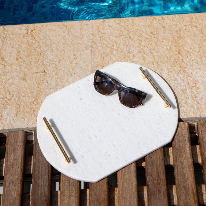 White oval capsule-shaped marble tray with brass handles placed by the pool